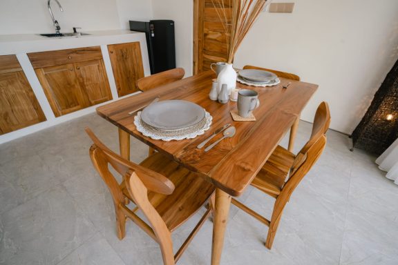 Table en bois avec de la vaisselle, chaises et décor minimaliste. Cuisine en arrière-plan.