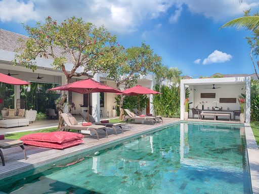 Villa avec piscine, chaises longues et parasols, entourée de verdure sous un ciel bleu.