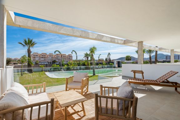 Terrasse avec chaises, vue sur piscine et palmiers sous un ciel bleu.