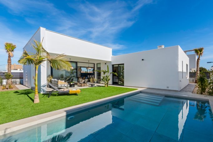 Villa moderne blanche avec piscine, pelouse et palmier sous un ciel bleu.