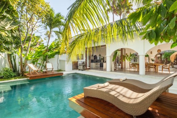 Piscine avec transats au bord, entourée de palmiers et d'une villa lumineuse.