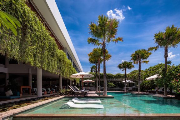 Vue d'une piscine entourée de palmiers et de chaises longues sous un ciel ensoleillé.